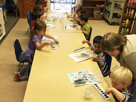 Preschool children using watercolors at a table with a teacher interacting with one of the children.